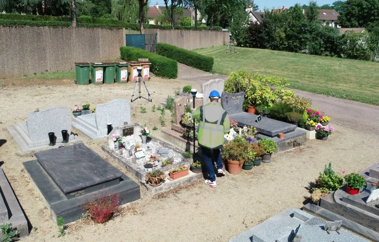 Un technicien Carteme en gilet haute visibilité relève un cimetière à pied, tombe par tombe ; une station totale sur trépied est installée en arrière-plan.